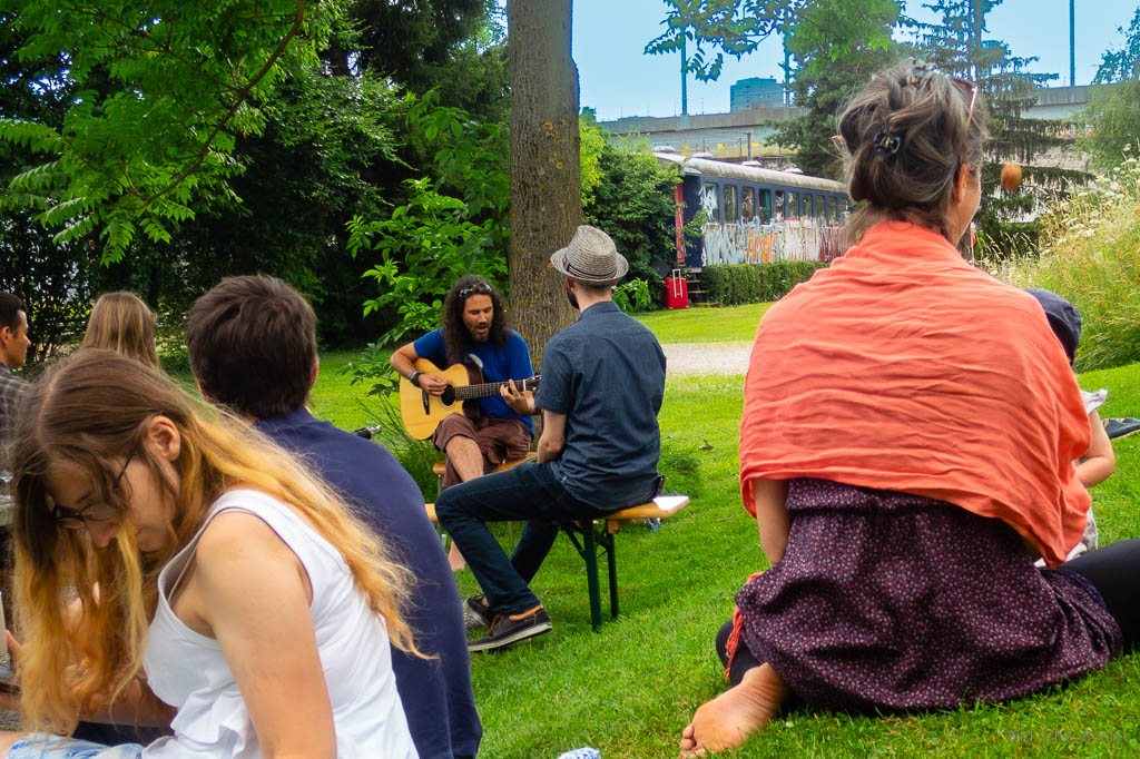 Eli Elk während Konzert vor Baum. Publikum auf Bänken und auf Gras am Hügel.